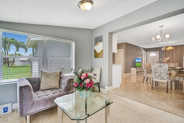 A living room with a grey sofa, a glass coffee table, and a vase of flowers. There is a dining area with patterned chairs and a chandelier.