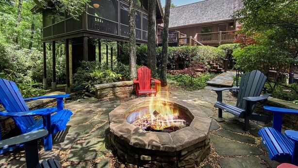 A stone fire pit surrounded by Adirondack chairs, a wooden deck, and a large window overlooking a lush garden.