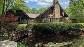 A rustic lodge with a stone chimney, wooden deck, and a view of the surrounding forest.