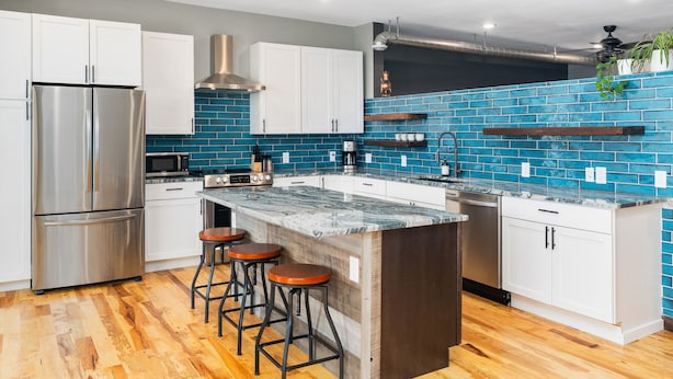A modern kitchen with white cabinets, a stainless steel refrigerator, a built-in oven, and a blue-tiled backsplash. - Craft District Loft - Michigan (Duluth)