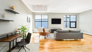 A modern living room with a wooden floor, a grey sofa, a brown leather chair, a wooden desk, and a large window. - Craft District Loft - Michigan (Duluth)