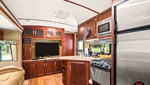 A compact RV kitchen with wooden cabinets, a stove, a refrigerator, and a flat-screen TV. - Fuel Your Passion for Epic Experiences and Outdoor Adventures at the Moto Ranch ATV! (Brooksville)