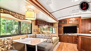 A compact RV interior with a dining area, seating, a TV, and wooden cabinets. - Fuel Your Passion for Epic Experiences and Outdoor Adventures at the Moto Ranch ATV! (Brooksville)