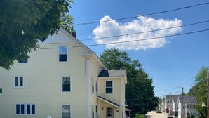 A two-story yellow house with a white picket fence, a driveway, and a street with other houses.