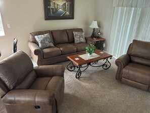 A living room with a brown leather sofa, two armchairs, a wooden coffee table with a plant, a lamp, and a clock on a side table.