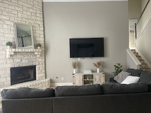 A living room with a stone fireplace, a flat-screen TV mounted on the wall, a gray sofa, and a staircase to the right. - Beautiful  Home with pool walking distance to Lake Austin 4 bedrooms  (Austin)
