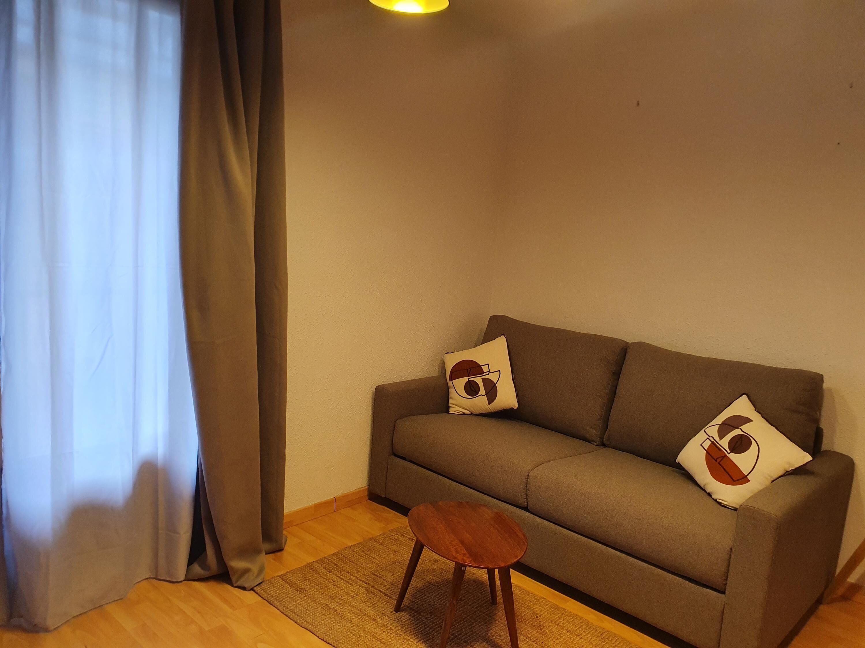 A cozy living room with a sofa, two decorative pillows, a small wooden stool, and a window with curtains.