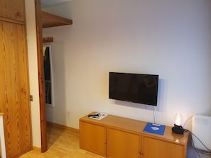 Una habitación con un armario de madera, un televisor montado y un escritorio con una lámpara y papeles.