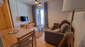 Een woonkamer met een bank, een aan de muur bevestigde televisie, een houten eettafel met stoelen en een kleine ronde tafel.