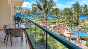 Een balkon met een glazen tafel en stoelen, met uitzicht op het zwembad van een resort, palmbomen en de oceaan.