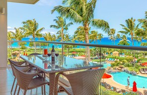 A glass-top table with wicker chairs, a bottle of wine, and two glasses on a balcony overlooking a pool and beach.