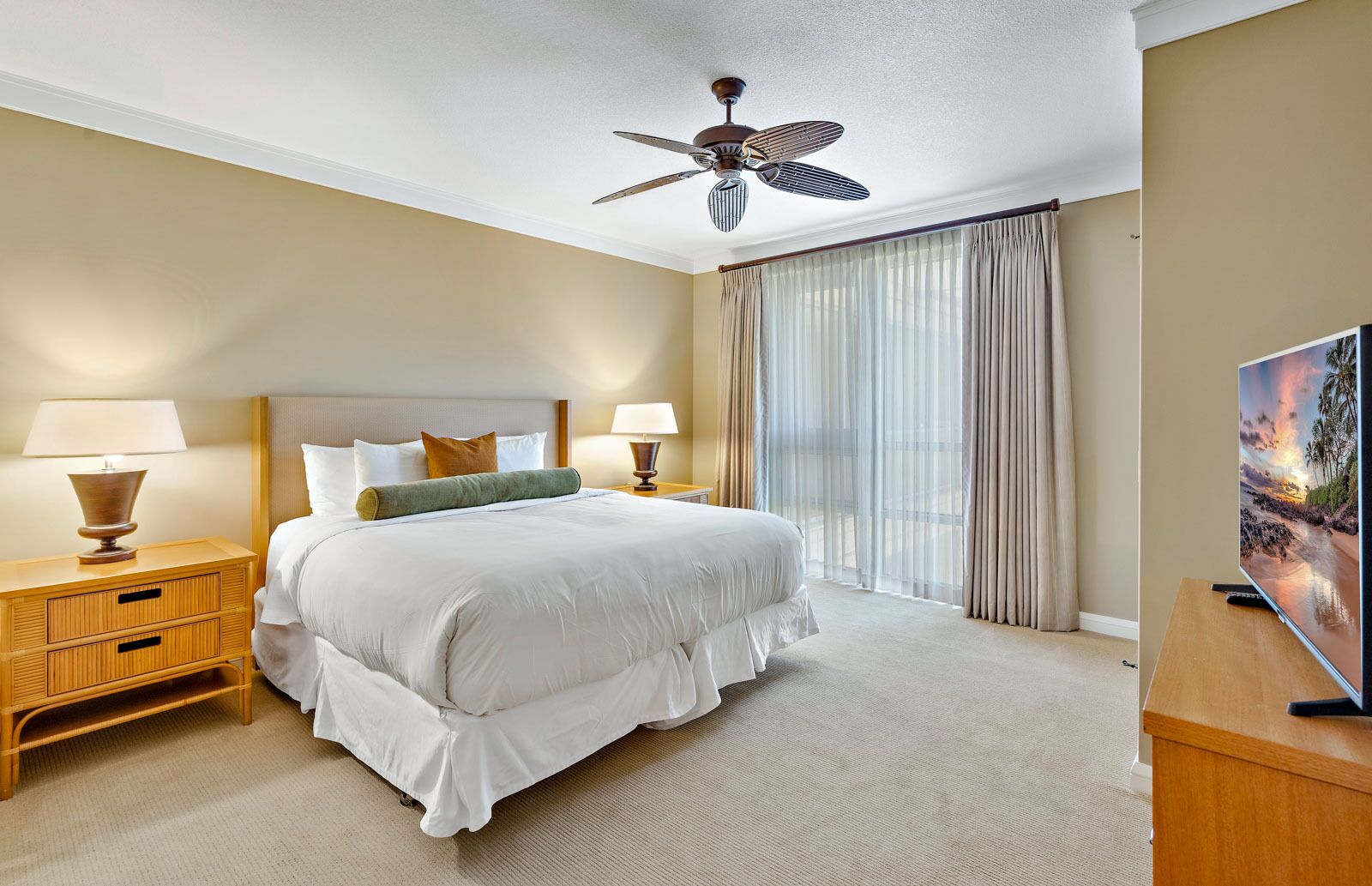 Une chambre d’hôtel avec un grand lit, deux tables de chevet avec des lampes, un ventilateur de plafond, une télévision et une grande fenêtre avec des rideaux.