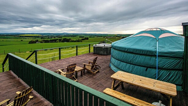 A wooden deck with a view of green fields and a cloudy sky. There is a large blue tent and a table with chairs.