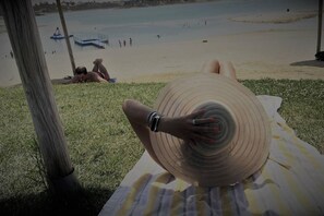 Eine Person liegt auf einem Strandtuch, trägt einen großen Hut und blickt auf einen Strand mit Menschen und Booten in der Ferne.