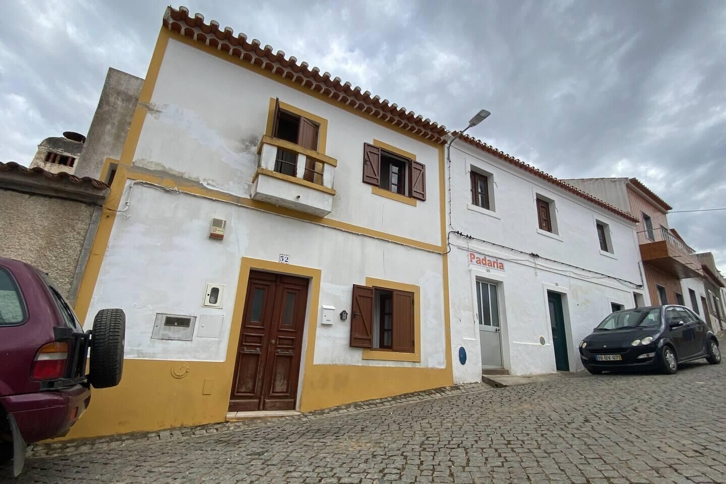 A two-story building with a sign reading "Padaria" on the ground floor, a brown door, and a cobblestone street.
