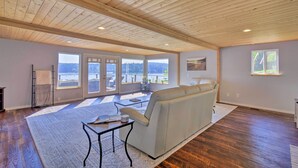 A spacious living room with a large sofa, a coffee table, and a view of the outdoors through large windows. - Waterfront Gig Harbor Home w/ Furnished Deck (Gig Harbor)