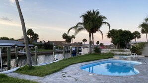 A swimming pool with a stone deck, surrounded by grass and palm trees, with a canal and boats in the background.