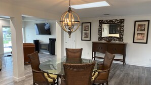 A dining area with a glass table and wicker chairs, a chandelier, and a fireplace in the background.