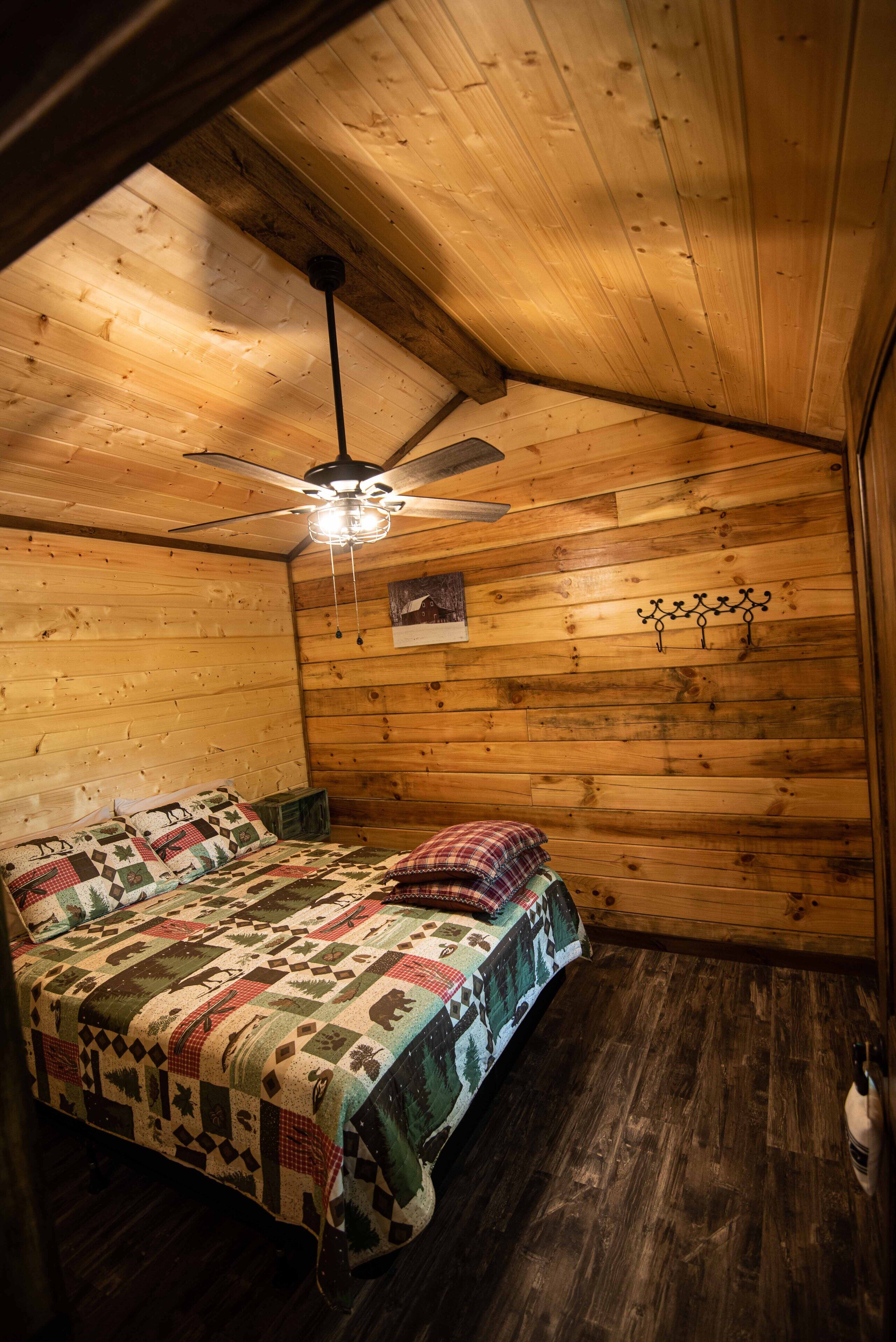 A cozy cabin room with a wooden ceiling, a bed with a quilt, a ceiling fan, and a small wall-mounted picture.
