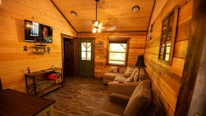 A cozy cabin interior with a wooden floor, a sofa, a television mounted on the wall, and a ceiling fan. - The Gilead Log Cabin (Harriet)