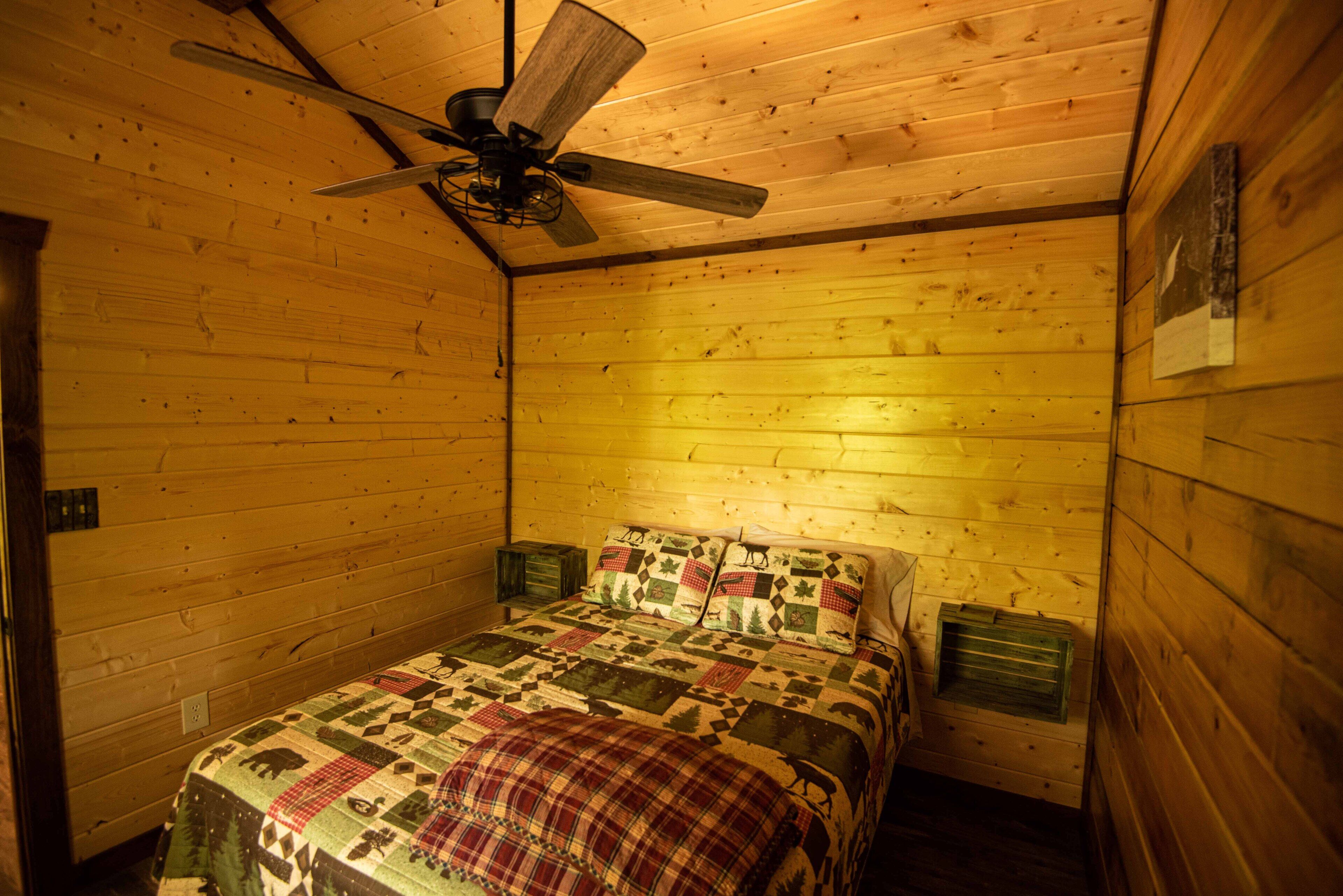 A wooden cabin bedroom with a bed, a ceiling fan, and a small window.