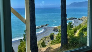 Vista costera a través de una ventana con vigas de madera, que muestra una playa, el océano y montañas distantes.