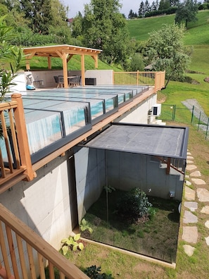A swimming pool with a wooden deck and a pergola, surrounded by a garden and a grassy hill.
