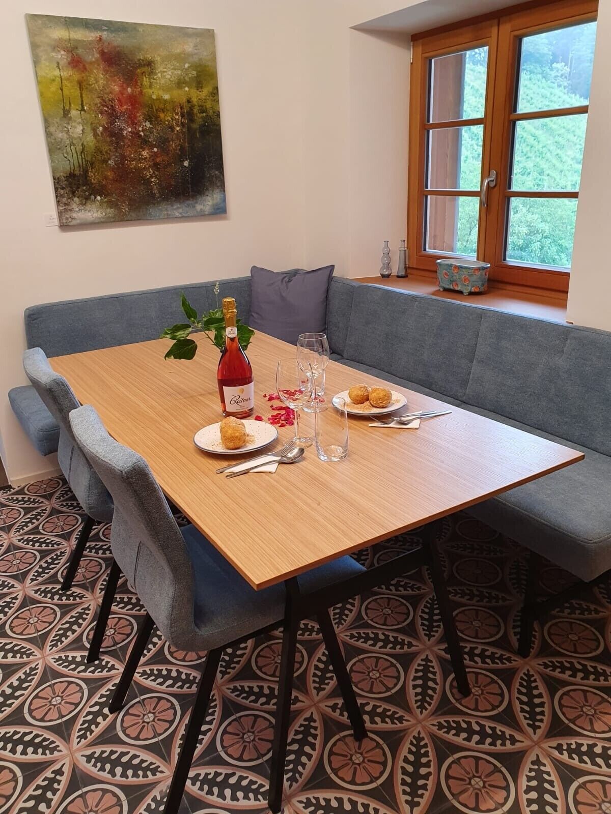 A dining area with a wooden table, grey upholstered sofa, patterned floor tiles, a bottle of red wine, and a painting on the wall.