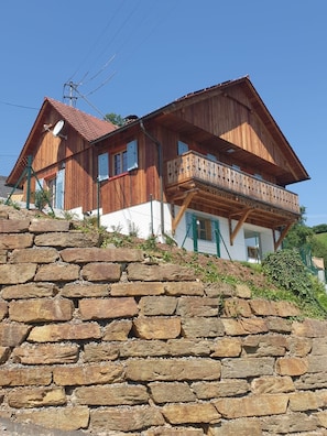 A wooden chalet with a stone retaining wall, a balcony, and a satellite dish.
