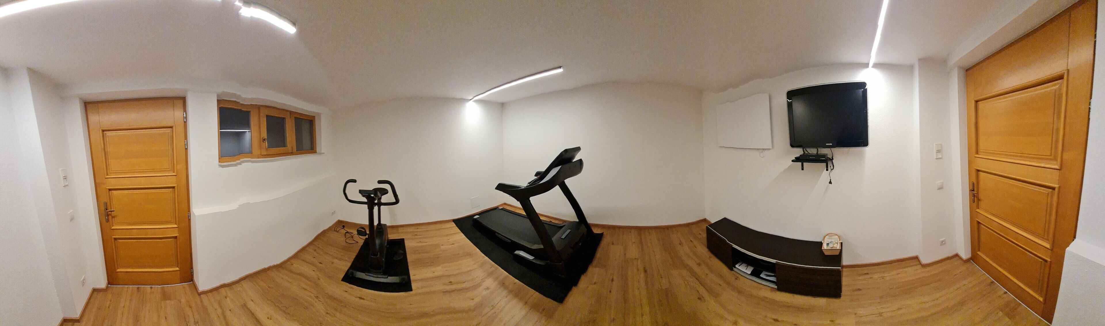 Una habitación con una cinta de correr, una elíptica, un televisor y armarios de madera.
