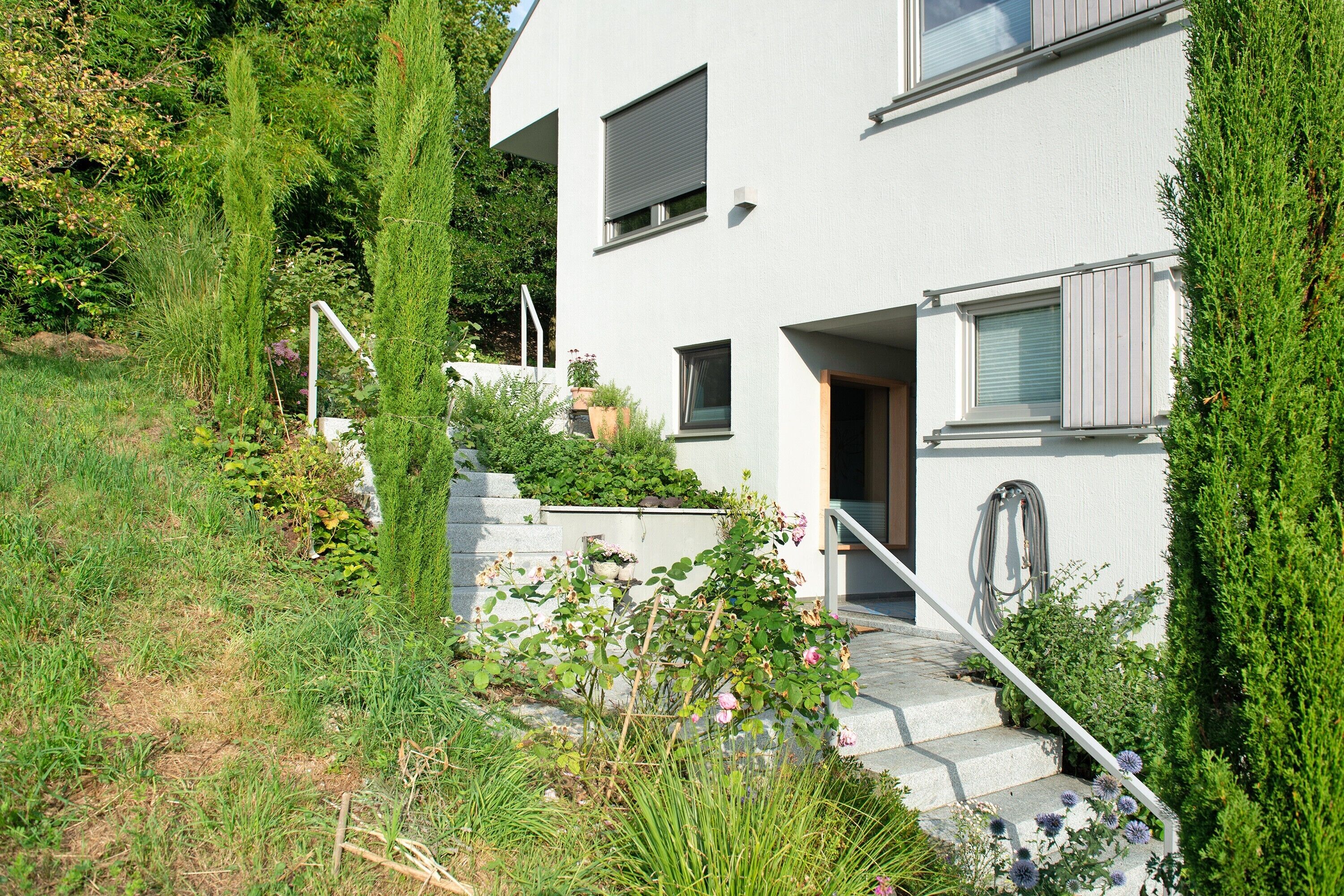 A modern two-story house with a stone staircase leading to the entrance, surrounded by lush greenery and a well-maintained garden.