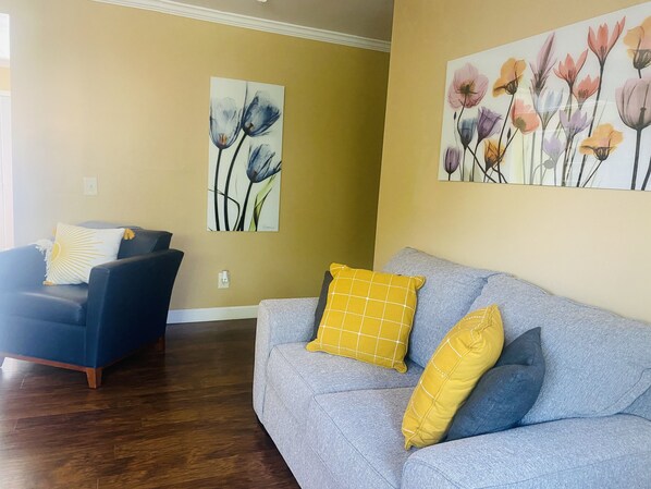 Una sala de estar con un sofá azul, un sillón gris y almohadas amarillas. Hay dos cuadros con motivos florales en las paredes.
