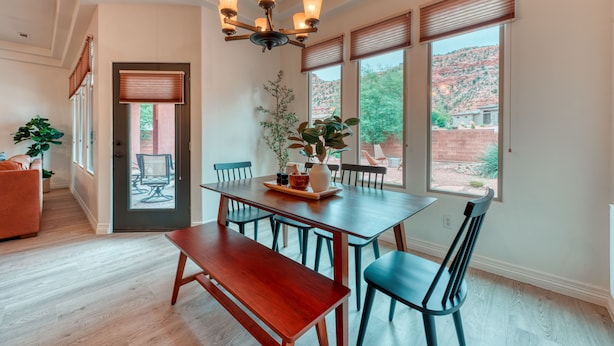 A dining area with a wooden table and chairs, a chandelier, and a view of a red rock landscape through large windows. - Red cliff views! 2 Kings! Private hot tub! Pool! (Kanab)