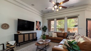 A living room with a large flat-screen TV, a ceiling fan, a sofa, a coffee table, and a window with a view of the outdoors. - Red cliff views! 2 Kings! Private hot tub! Pool! (Kanab)