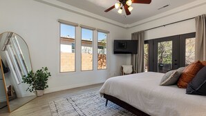 A bedroom with a large bed, a ceiling fan, a TV, and a view of the outside. - Red cliff views! 2 Kings! Private hot tub! Pool! (Kanab)