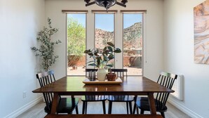 A dining area with a wooden table, black chairs, a potted plant, and a view of a mountainous landscape through large windows. - Red cliff views! 2 Kings! Private hot tub! Pool! (Kanab)