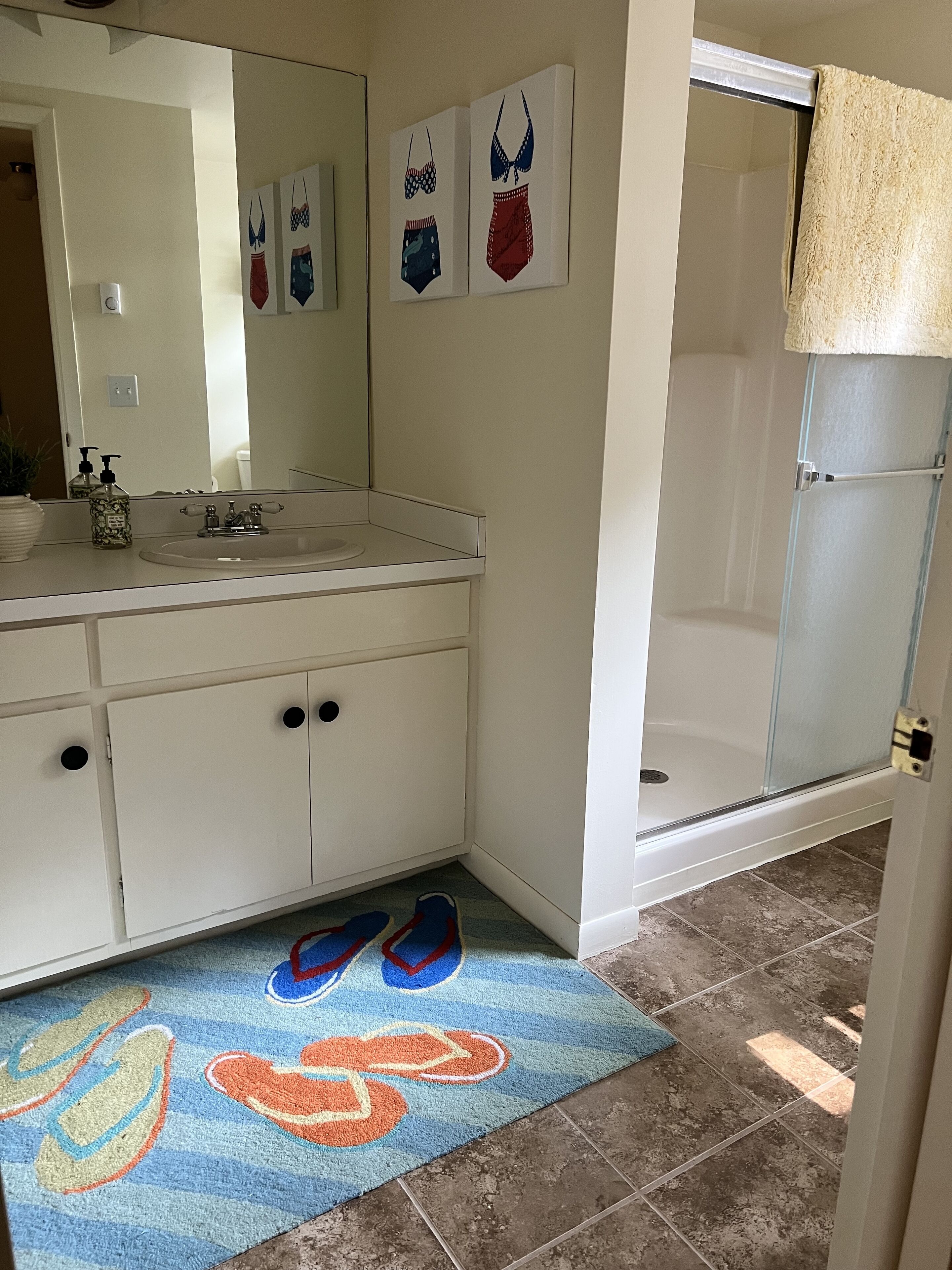Une salle de bain avec un lavabo blanc, un miroir et des armoires. Deux tableaux encadrés sont accrochés au mur, une paire de pantoufles est posée sur le sol et il y a une porte de douche.