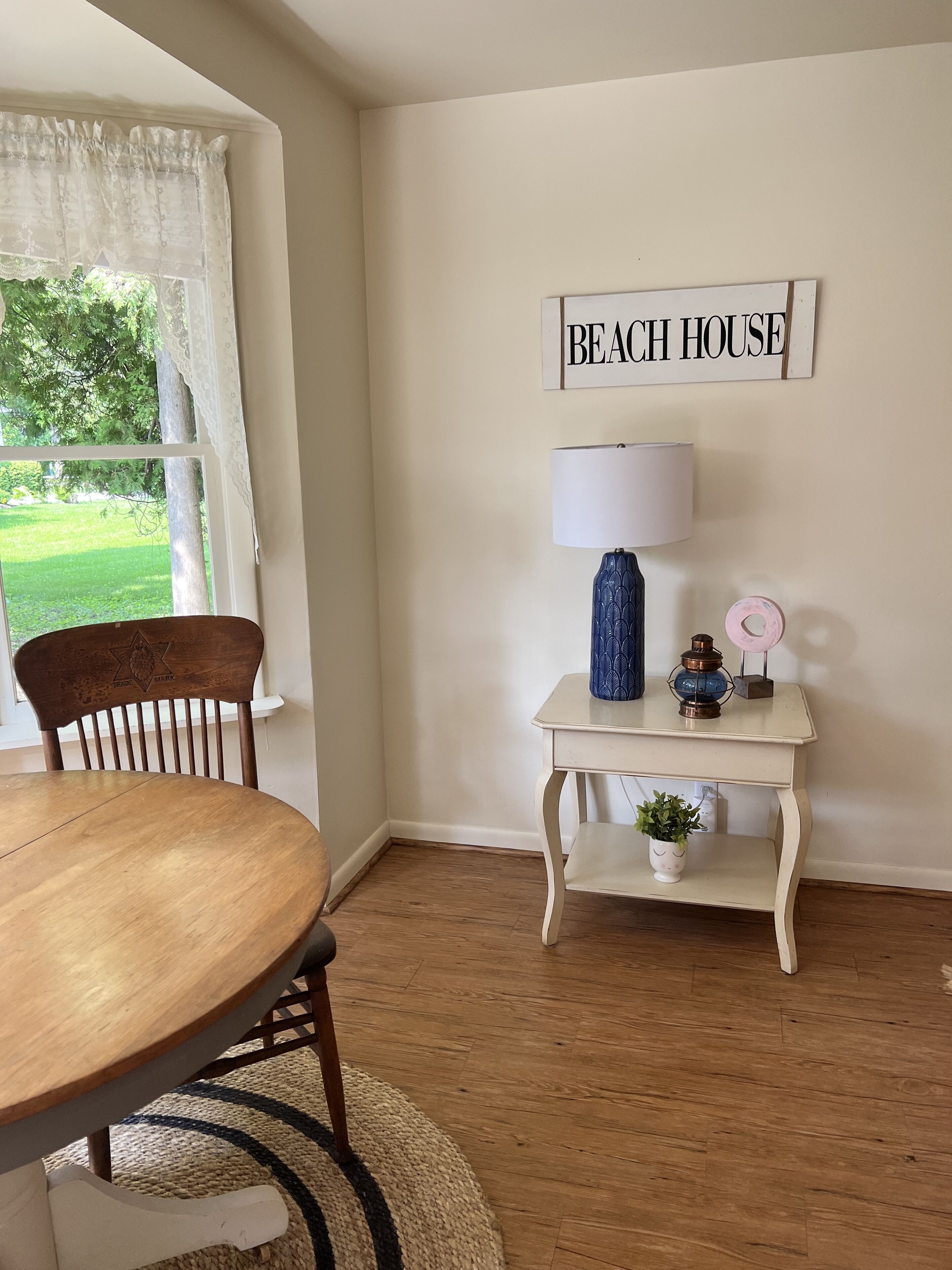 Un área de comedor con una mesa redonda de madera, una silla, una lámpara, una mesita con adornos y un letrero que dice "BEACH HOUSE".
