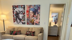 Ein Wohnzimmer mit einem beigefarbenen Sofa, einem Couchtisch und einem gemusterten Teppich. Darüber befinden sich zwei Wandbilder.