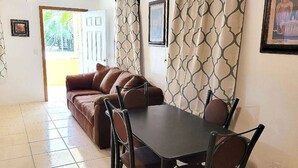 Ein Wohnzimmer mit braunem Sofa, einem Esstisch mit vier Stühlen, gemusterten Vorhängen und einer Tür, die nach draußen führt.