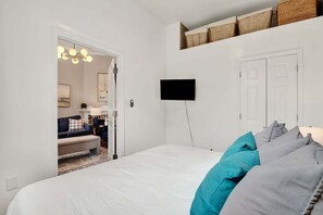A bedroom with a bed, a TV mounted on the wall, a door, and a small shelf with baskets.