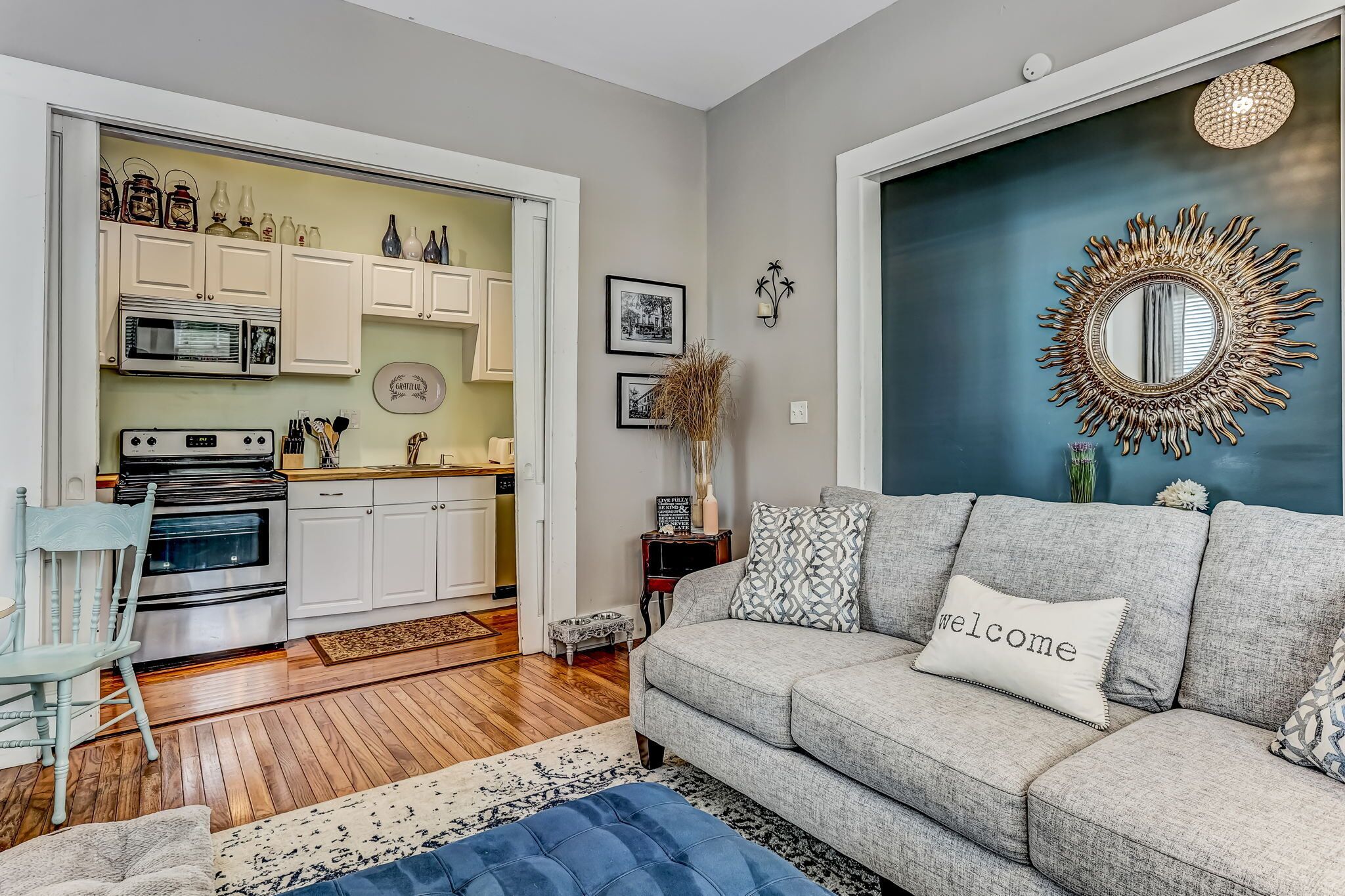 A modern living room with a grey sofa, a blue ottoman, and a 'welcome' pillow. A kitchen is visible in the background with white cabinets and a microwave.