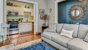Una sala de estar moderna con un sofá gris, un otoman azul y una almohada que dice 'welcome'. Se ve una cocina al fondo, con armarios blancos y un microondas.