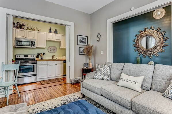 A modern living room with a grey sofa, a blue ottoman, and a 'welcome' pillow. A kitchen is visible in the background with white cabinets and a microwave. - Houston Street Retreat Just Steps from Crawford Square | Heated Pool Access (Savannah)