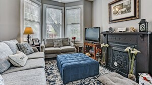 Una sala de estar con sofá, reposapiés, chimenea, televisor y obras de arte enmarcadas.