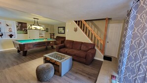 A living room with a pool table, a brown leather couch, a coffee table, and a staircase.