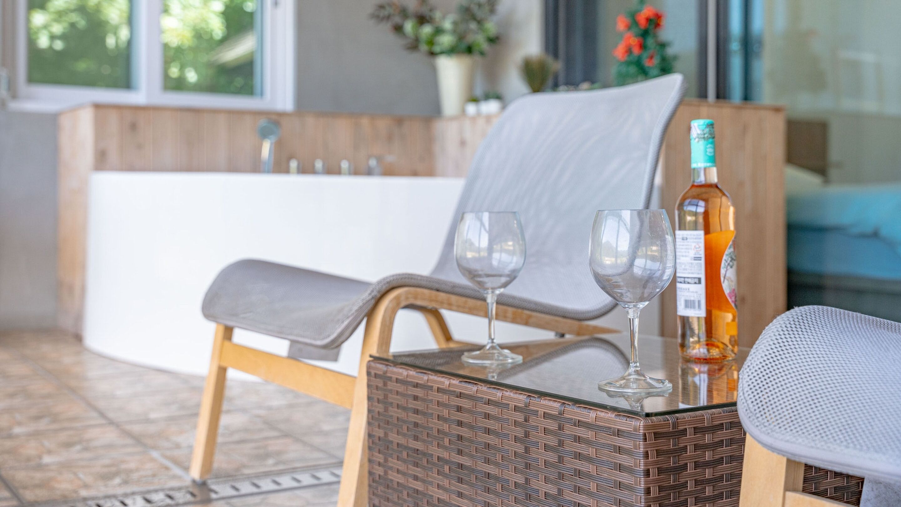 A wicker chair with a grey cushion, two empty wine glasses, and a bottle of liquor on a glass table.