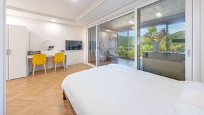 A bedroom with a large bed, a desk with two yellow chairs, a TV, and a sliding glass door leading to a balcony with a view of greenery. - Hongcheon Toliaigeullaemping (Hongcheon)