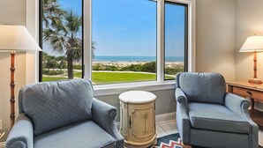 A room with two armchairs, a small table, and a window with a view of the beach and palm trees. - 2nd-Floor Turtle Dunes Unit with Breathtaking Views (Fernandina Beach)