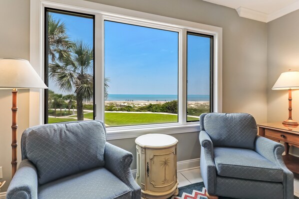 A room with two armchairs, a small table, and a window with a view of the beach and palm trees.